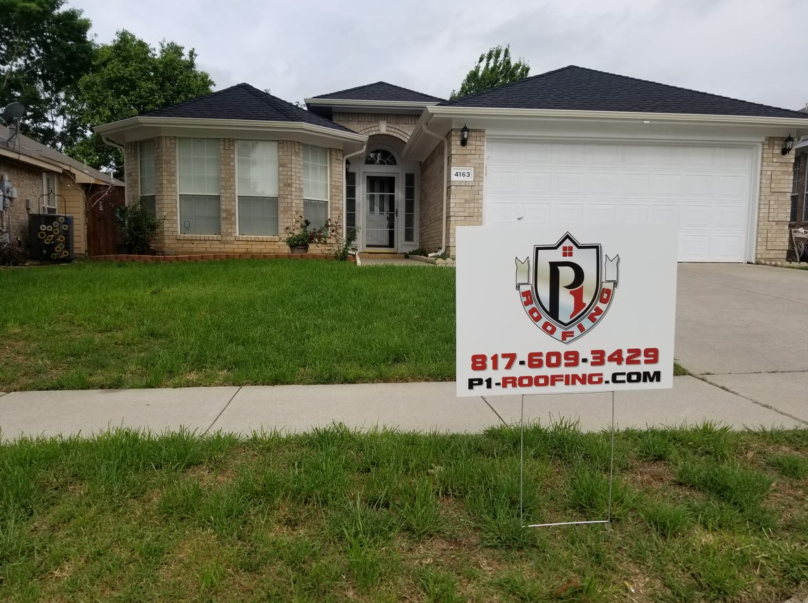 House with new black roof, lawn, and a roofing company sign in front.
