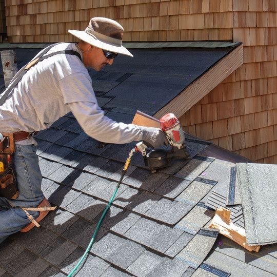 Roofer, wearing a hat, uses a nail gun to install asphalt shingles on a roof.