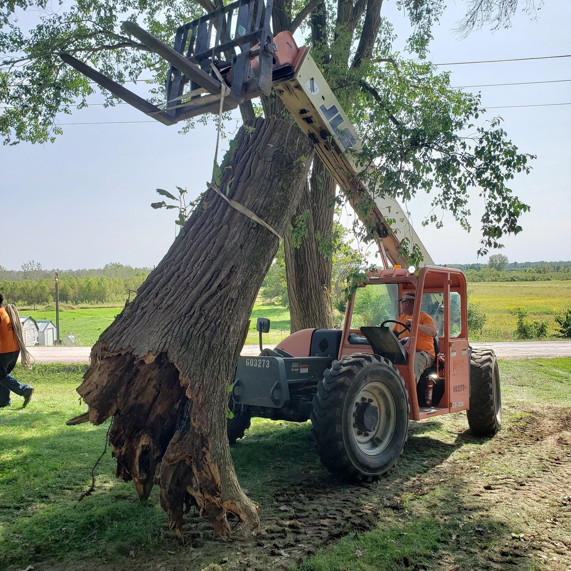A telehandler lifting a large tree trunk. A person in an orange vest walks on the left. Green field in background.
