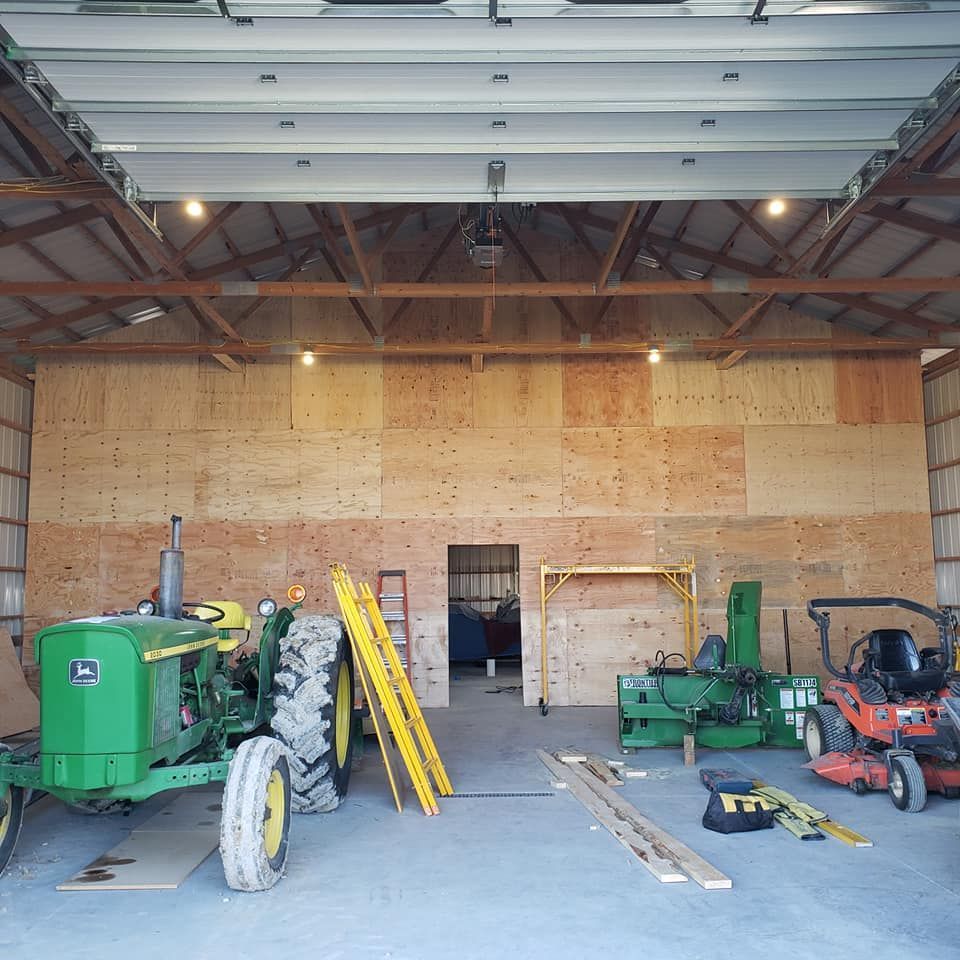 Inside a barn: a green tractor, construction materials, equipment, and a plywood-covered wall.