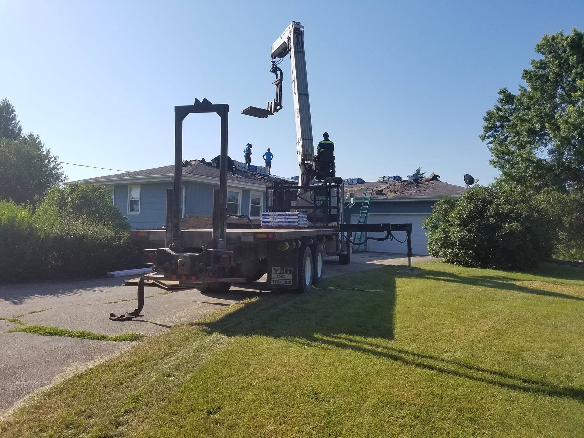 Roofers on a house with a crane placing supplies; sunny day, green grass.