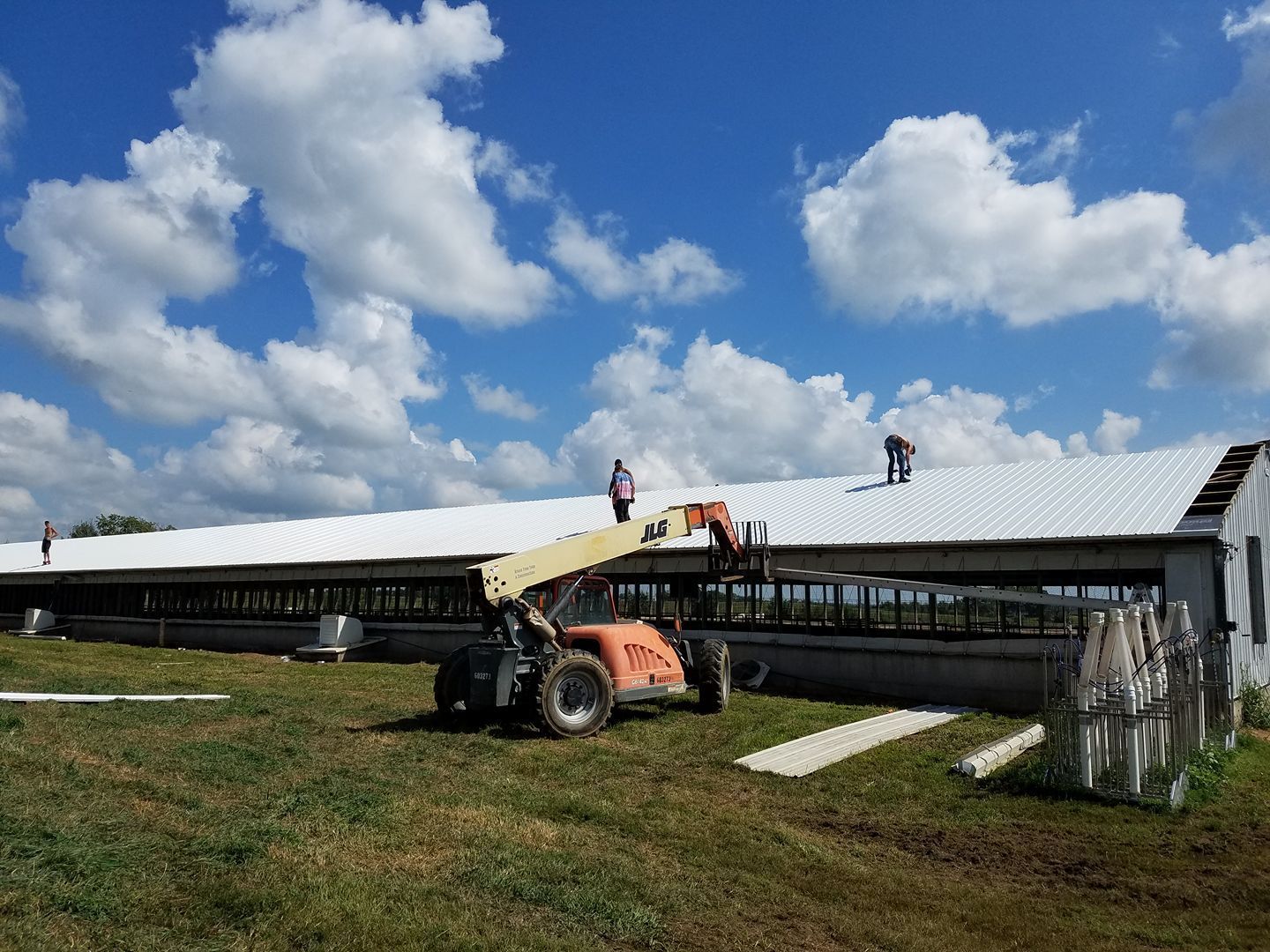 Construction workers on a roof, using a lift. Bright blue sky with clouds. White building in a field.