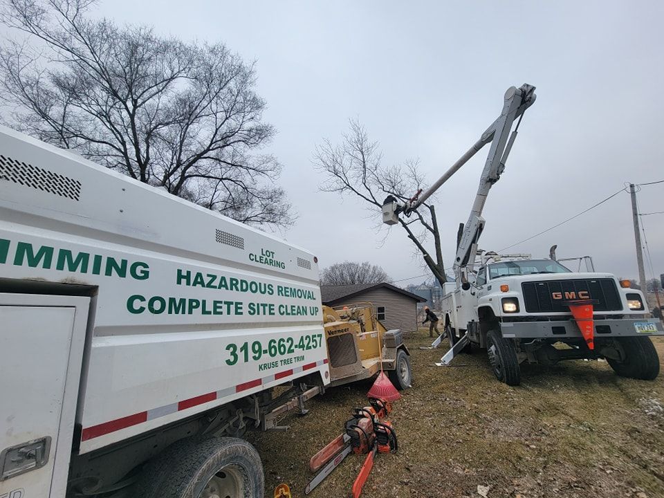 Tree removal service in progress: bucket truck, chipper, and truck, trimming tree branches in outdoor setting.