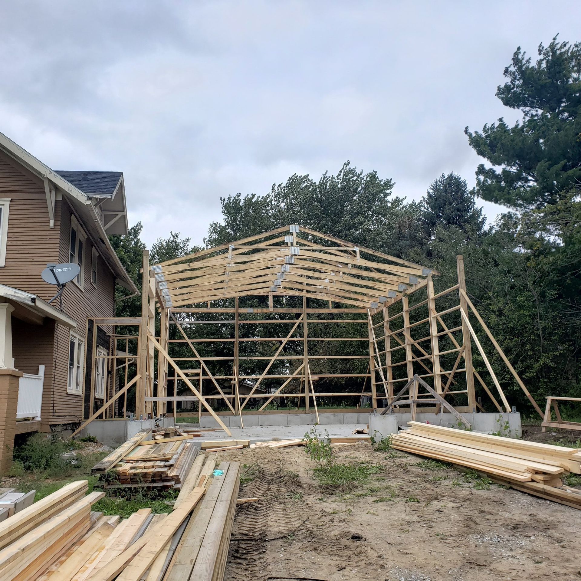 Construction site: wooden frame of a garage under cloudy sky next to a house. Lumber piles in foreground.