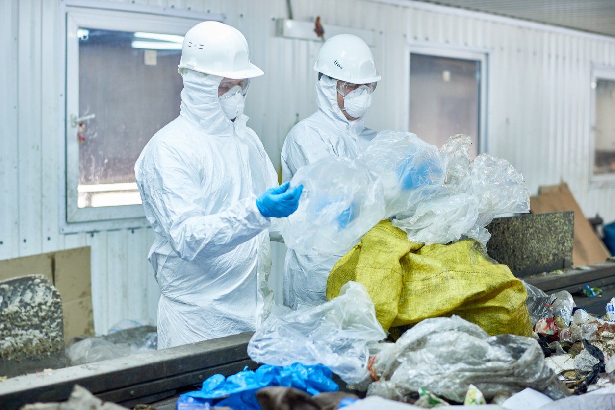 Two men in protective suits are standing next to a pile of plastic bags.
