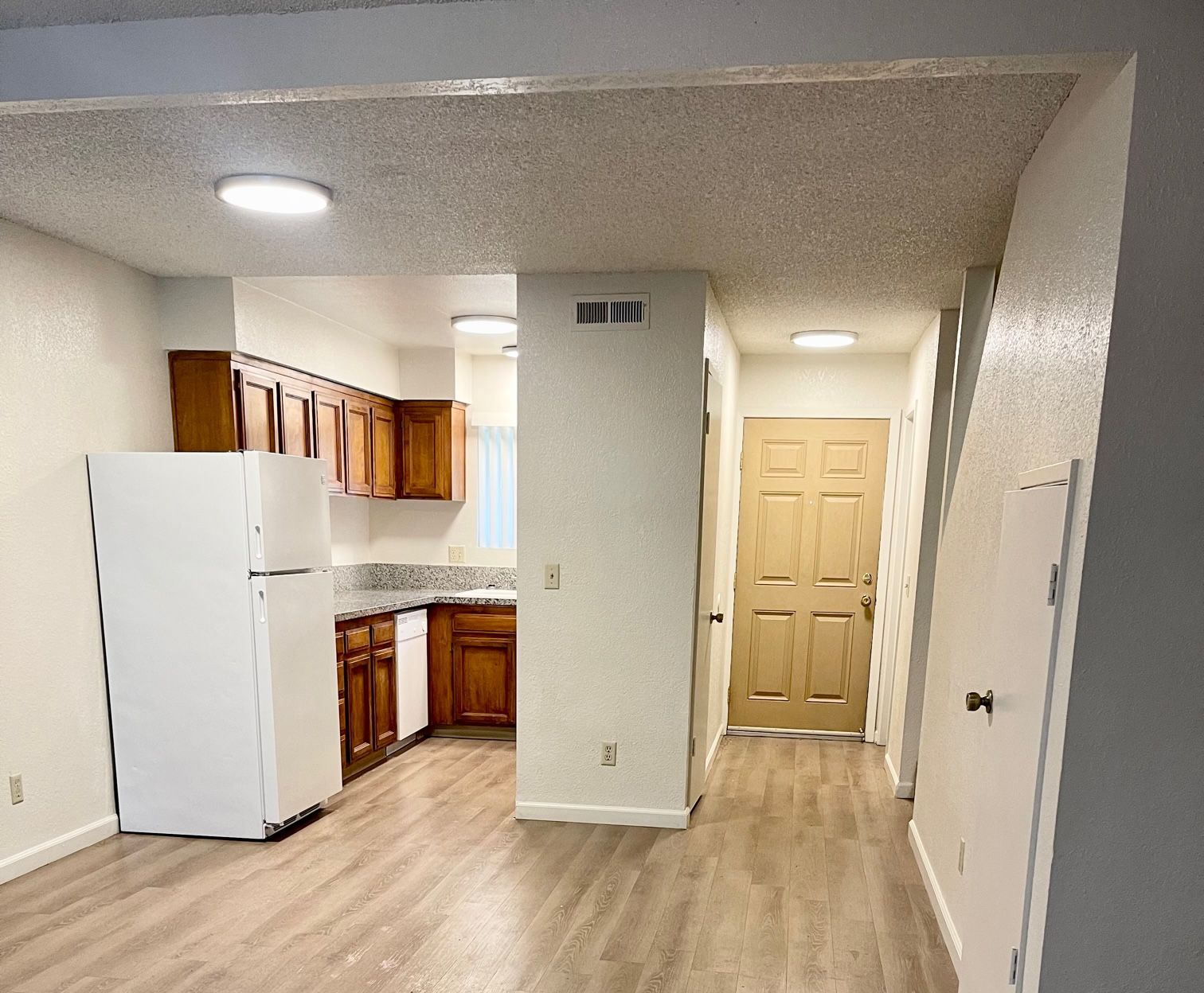 Open-concept apartment with a kitchen and entryway. Light wood cabinets, white fridge, and wood-look flooring.