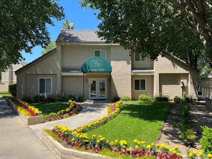 Beige two-story building with a green awning and double doors, surrounded by flower beds, grass, and trees.