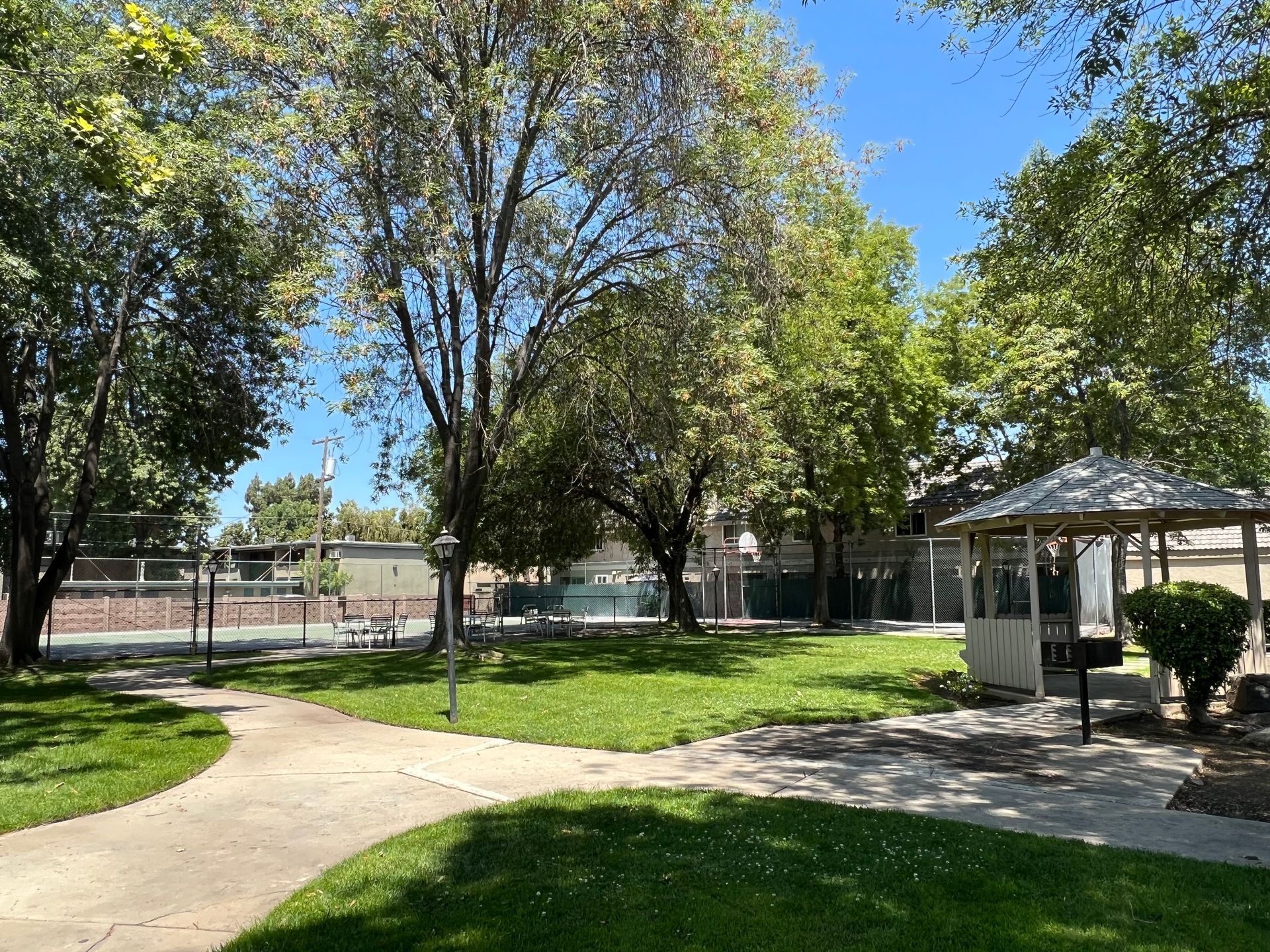 Park scene with grass, trees, picnic table, paved path, gazebo, and fence under a sunny sky.