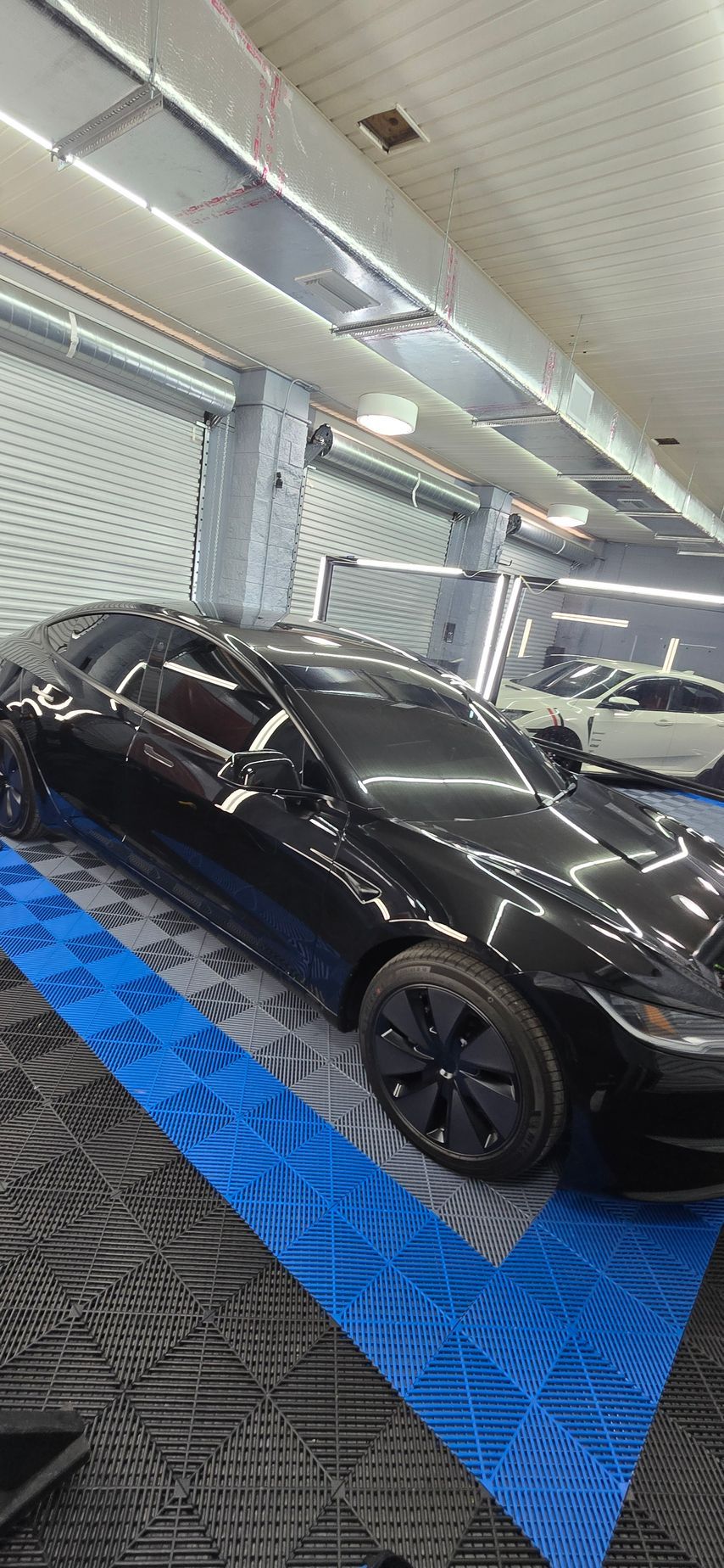 Black sedan on a blue display mat inside a showroom or garage, viewed from the front-left side