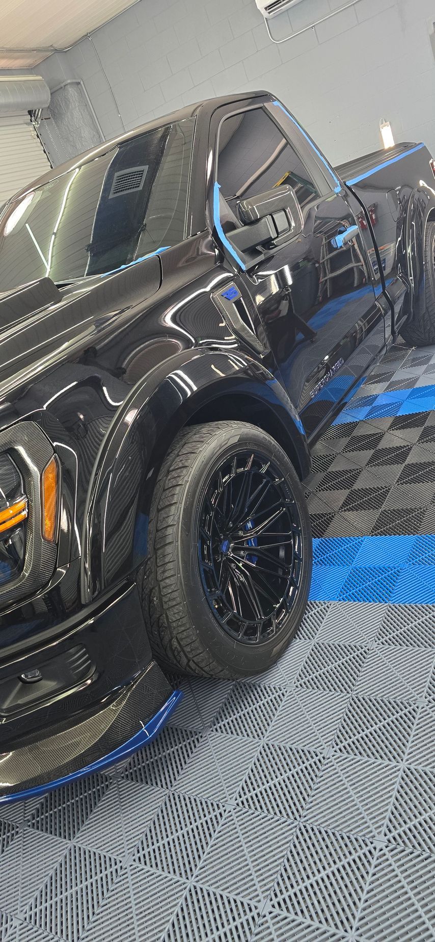 Black pickup truck with off-road tires parked indoors on a blue-and-gray checkered floor.