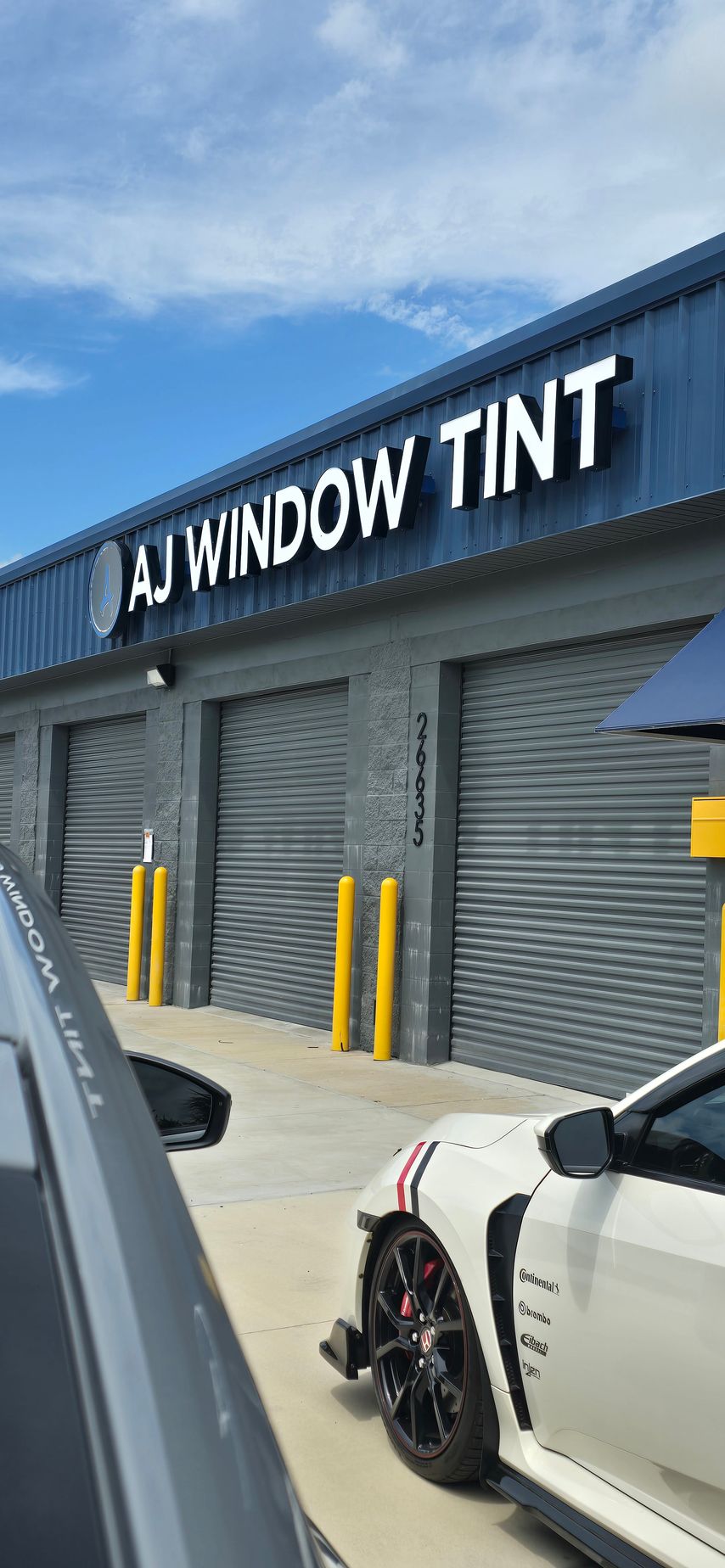 Storefront with “WINDOW TINT” sign, gray shutters, and parked cars outside under a blue sky