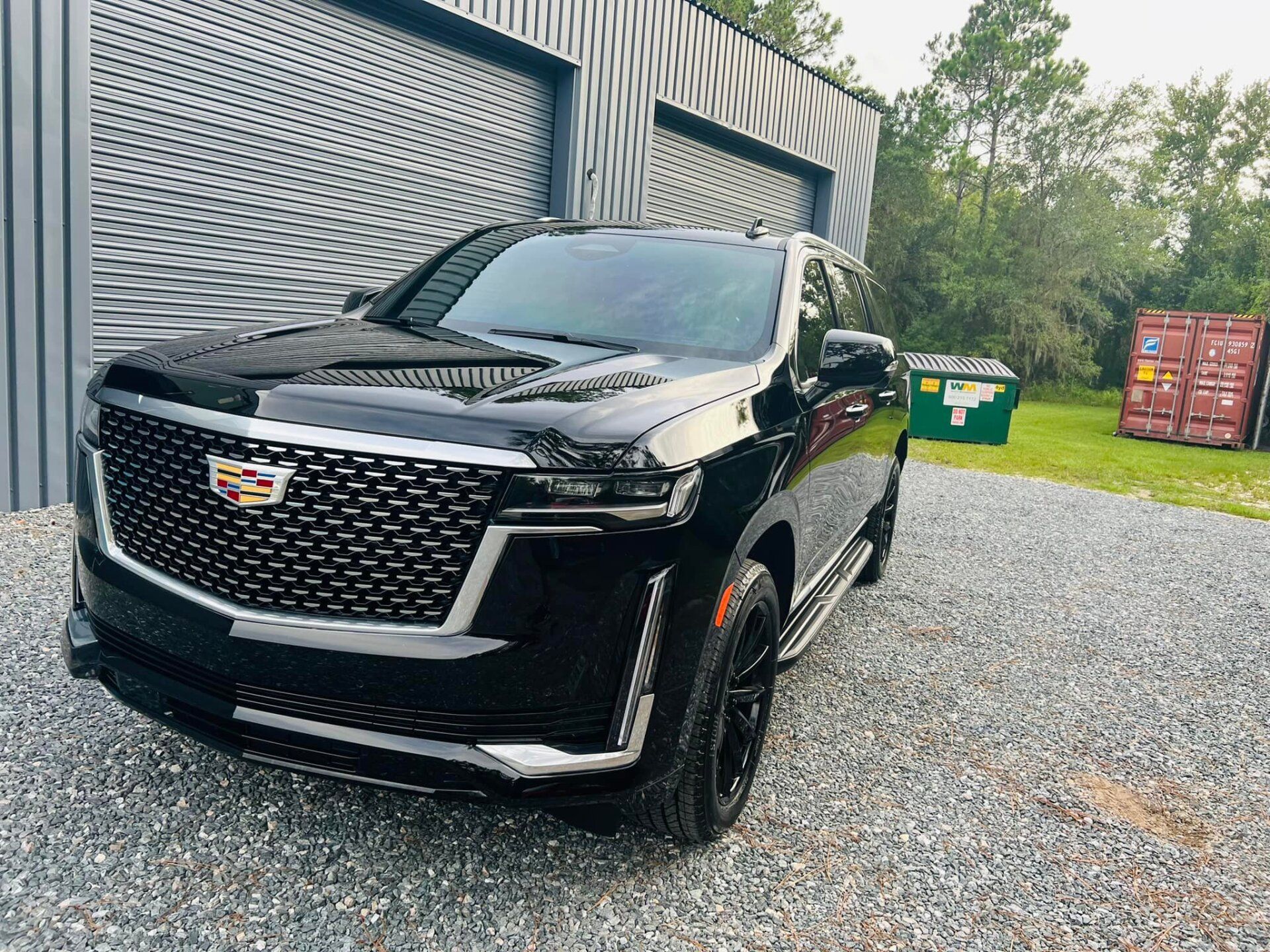 Black Cadillac Escalade SUV parked by a corrugated metal building with a trash bin and shipping container nearby.