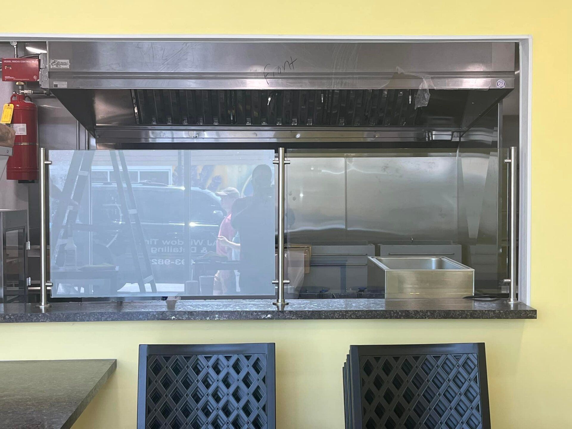 Restaurant pass-through with stainless steel hood, glass dividers, and dark patterned chairs below.
