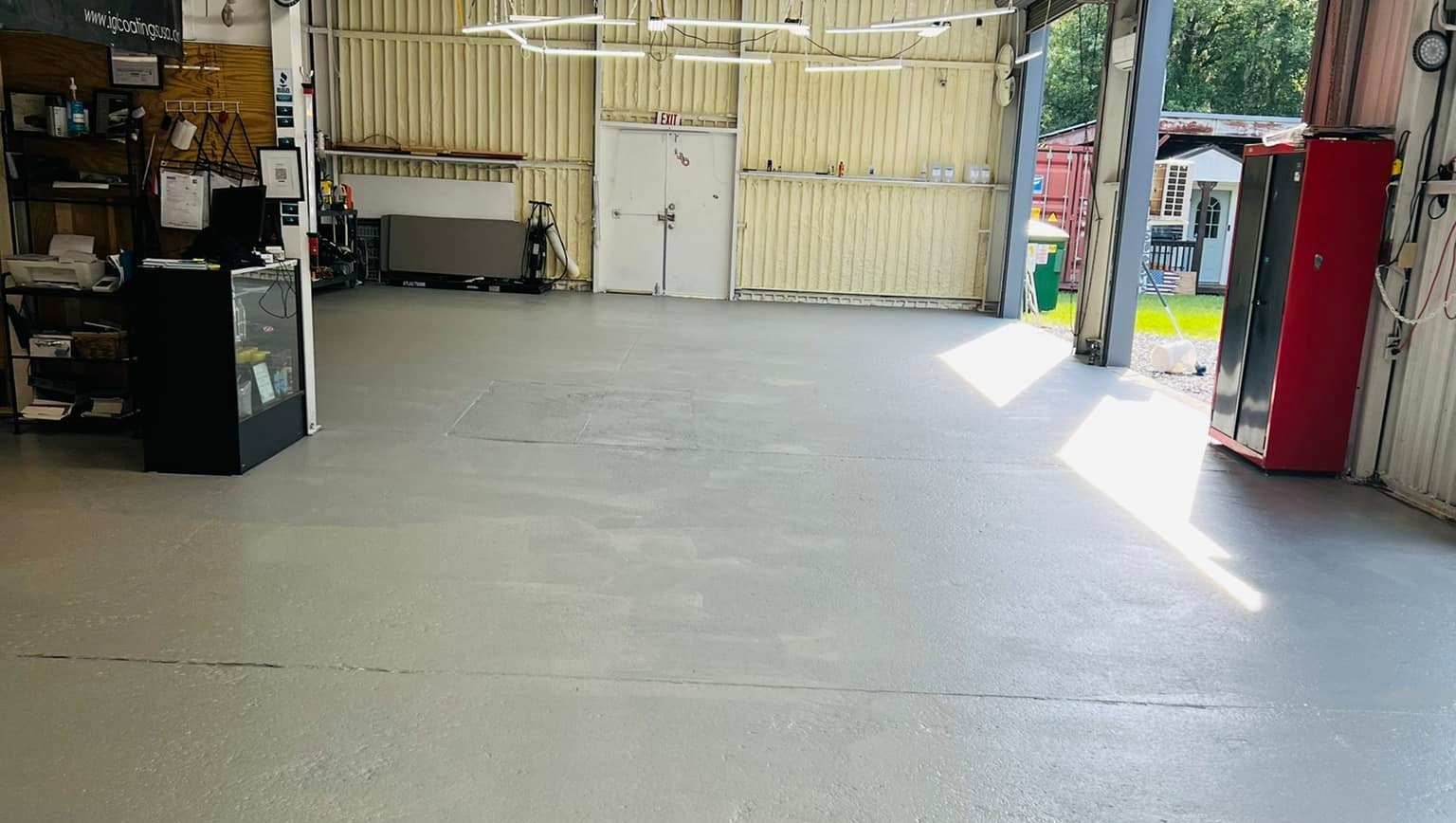 Empty gray-floored garage, bright sunlight streams in from an open doorway, a red cabinet on the right.