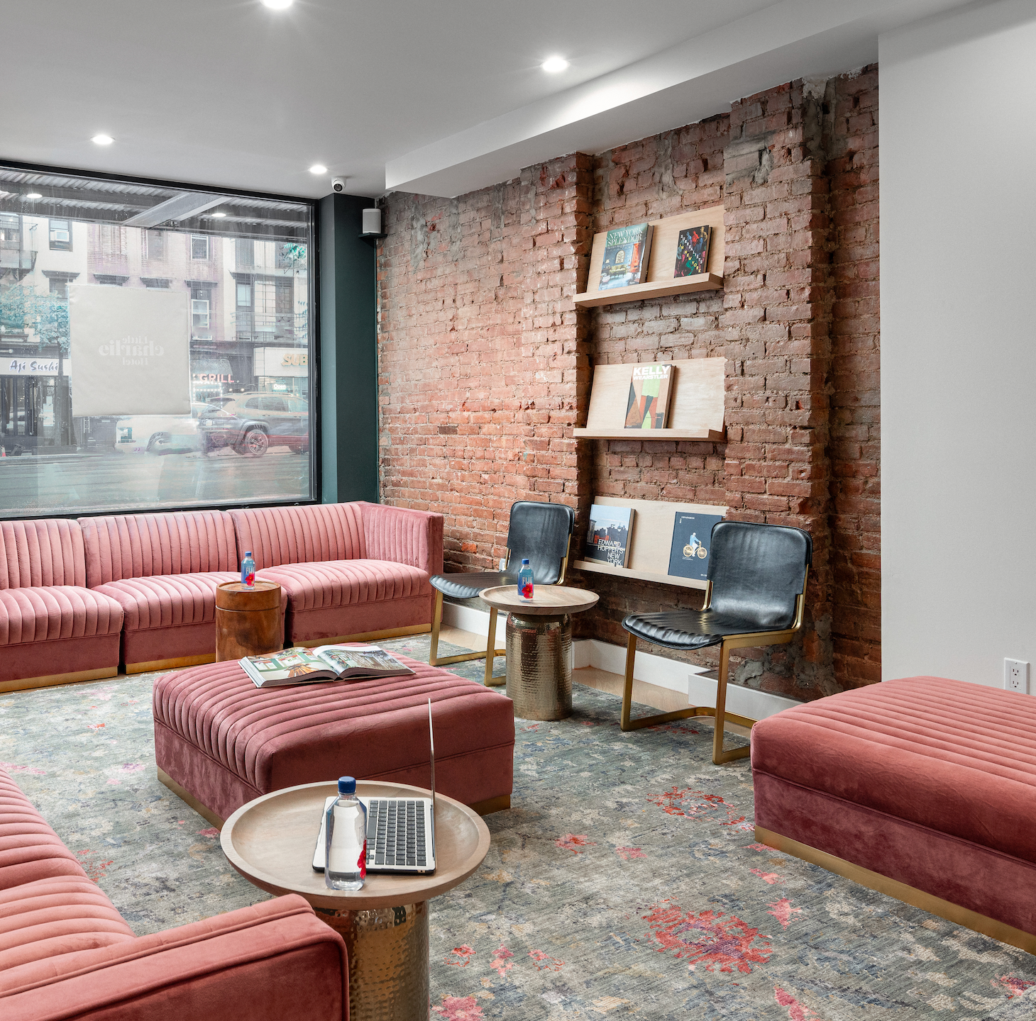 A waiting room with pink velvet furniture, exposed brick wall, and large window overlooking a city street.