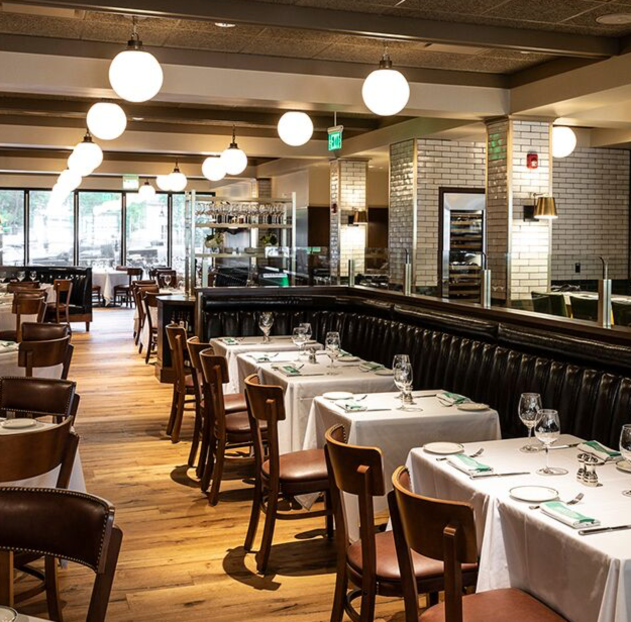 Restaurant interior with white-clothed tables, brown chairs, globe lights, and a partial view of the kitchen.