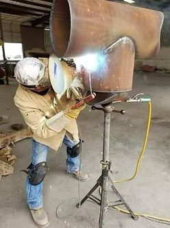 Welder in protective gear welding large metal pipe section, standing on a tripod in a workshop.
