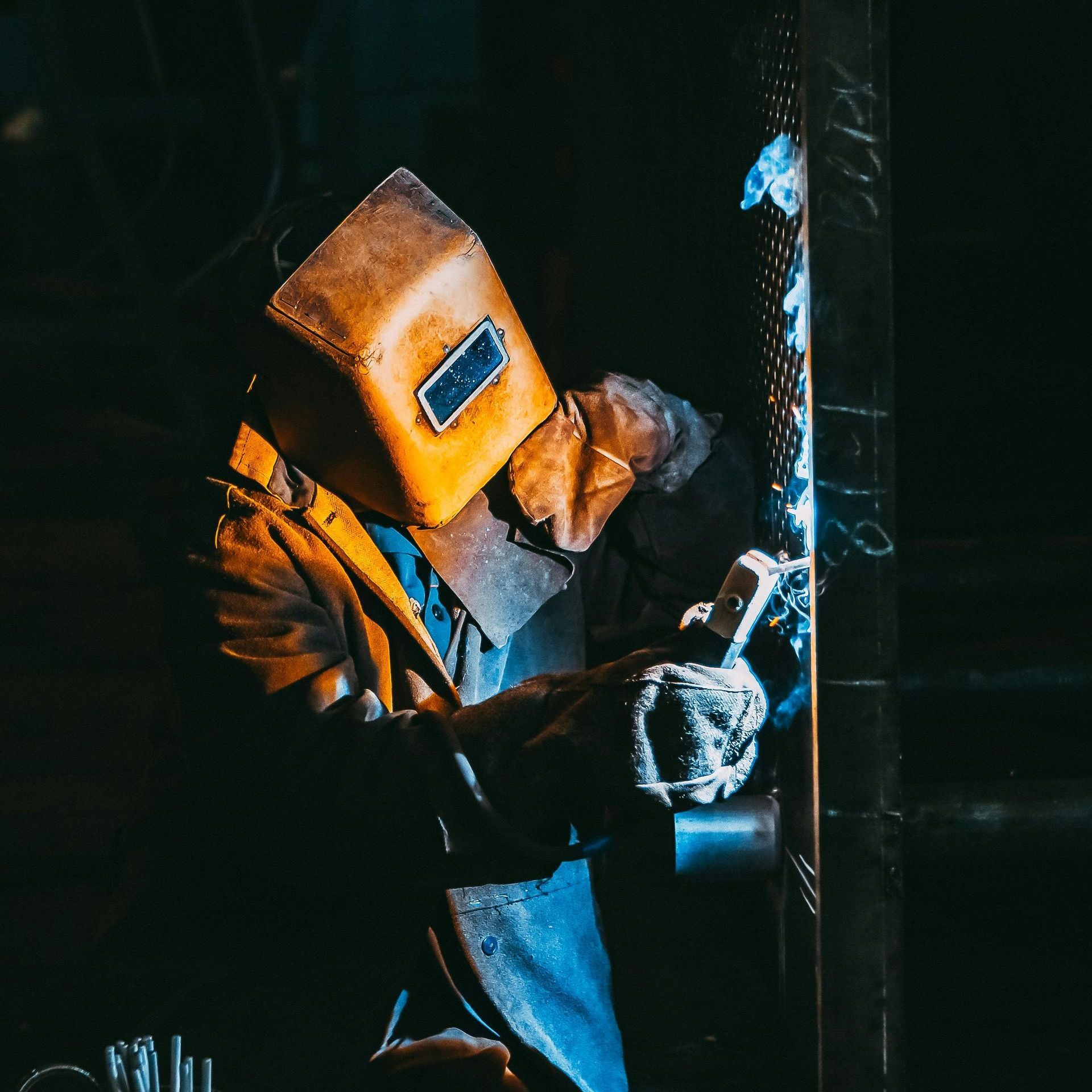 Welder in protective gear using a welding torch on metal in a dimly lit workshop.
