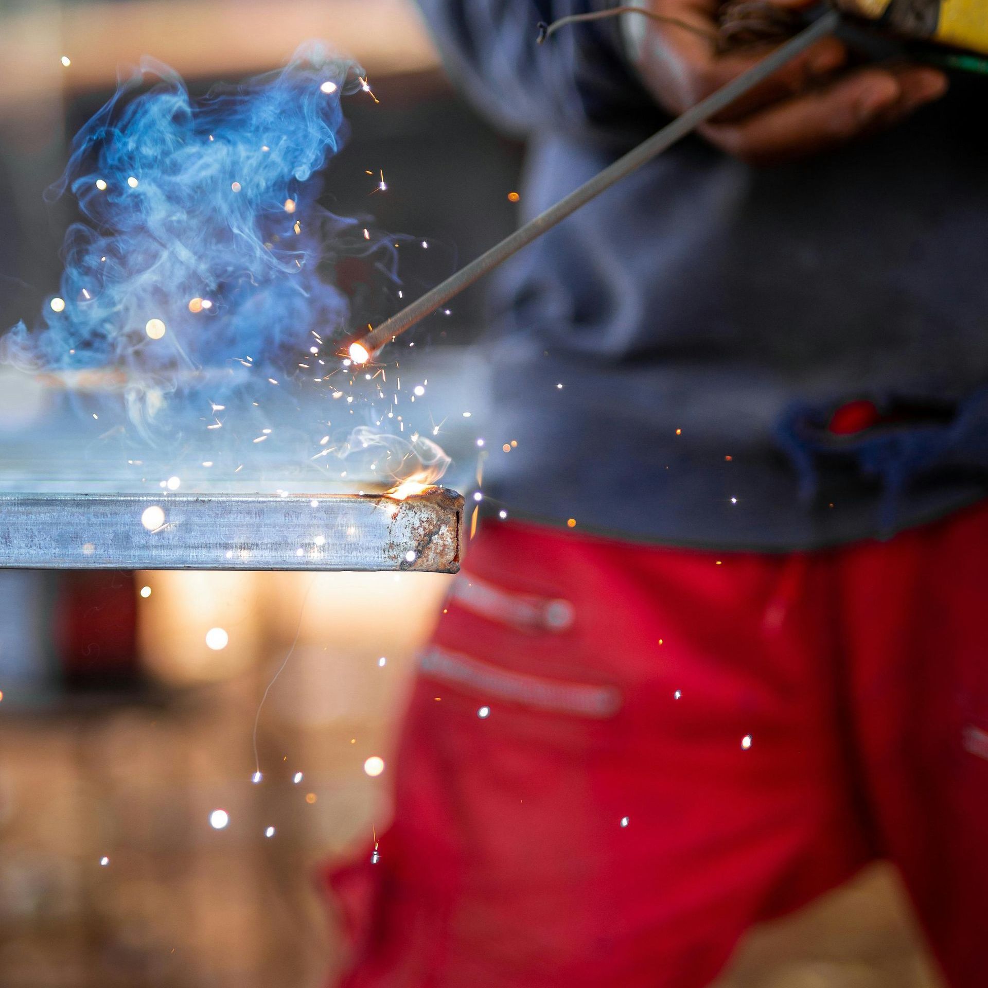 Welder in red pants working on metal, sparks and blue smoke.