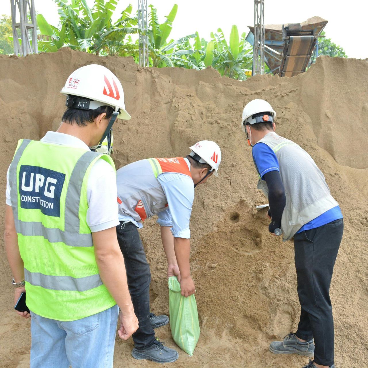 Three workers examining sand pile; one filling a bag.