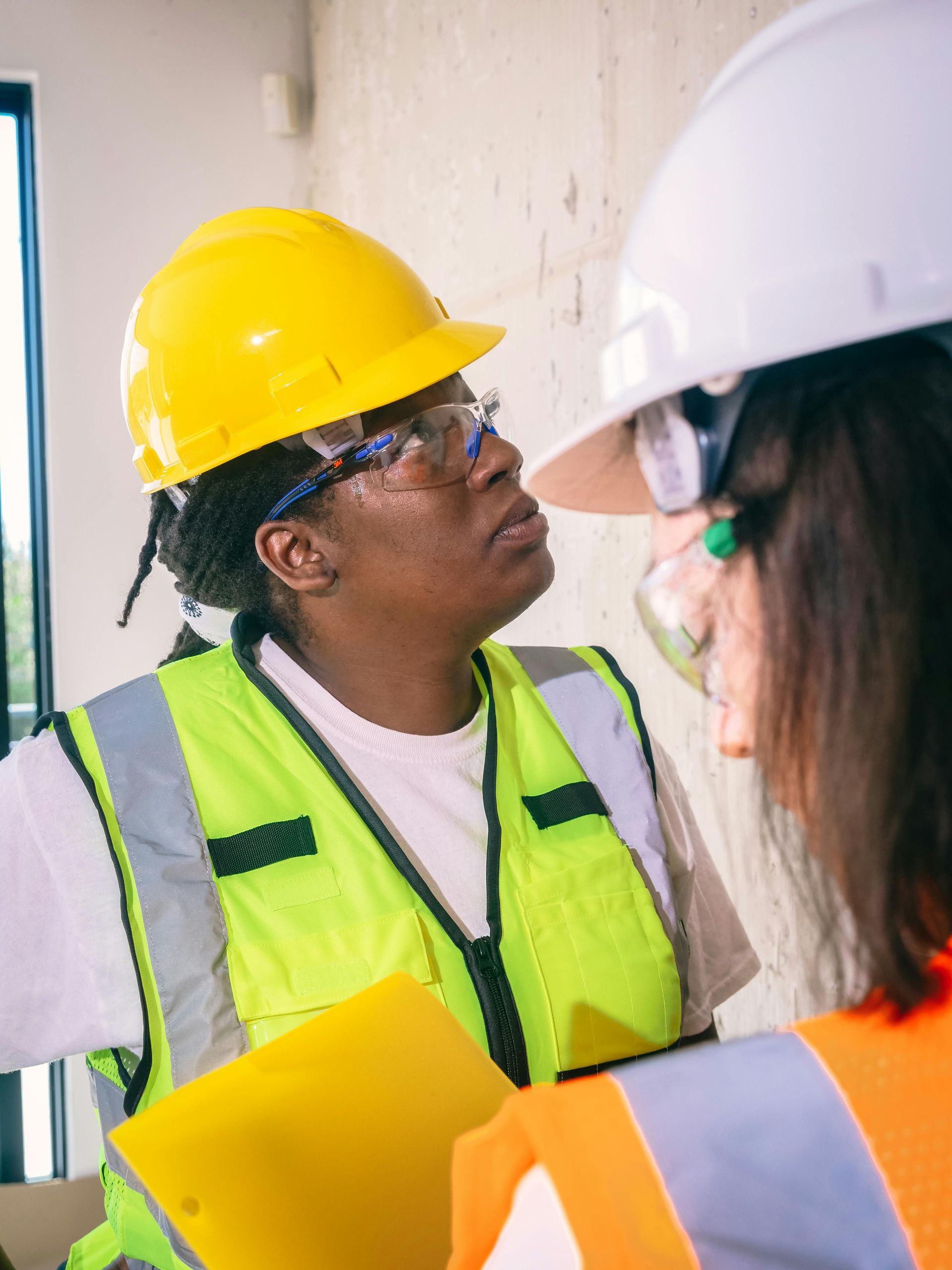 Two workers in safety gear talking on site. One in yellow hard hat, holding a clipboard.