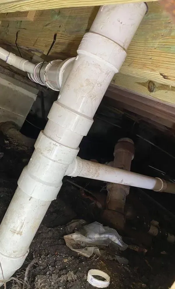 White PVC plumbing pipes under a wooden structure, with several joints and connections visible.