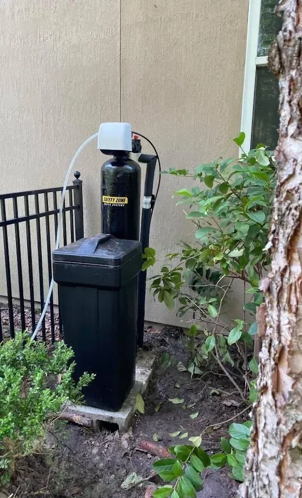 Black water softener system next to a fence and bushes in a residential yard.