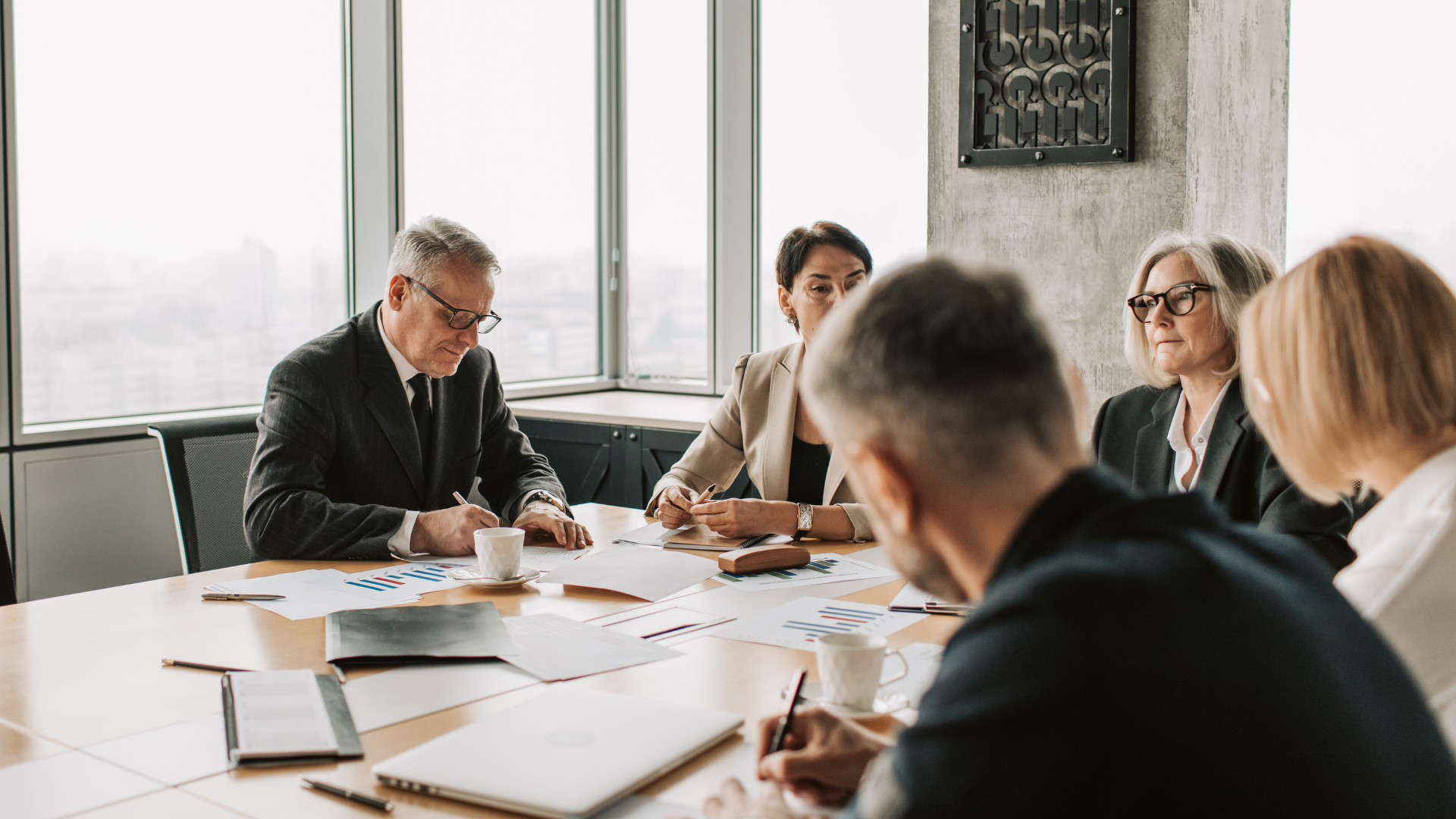 Business people in suits at a meeting, reviewing papers around a table in a modern office.