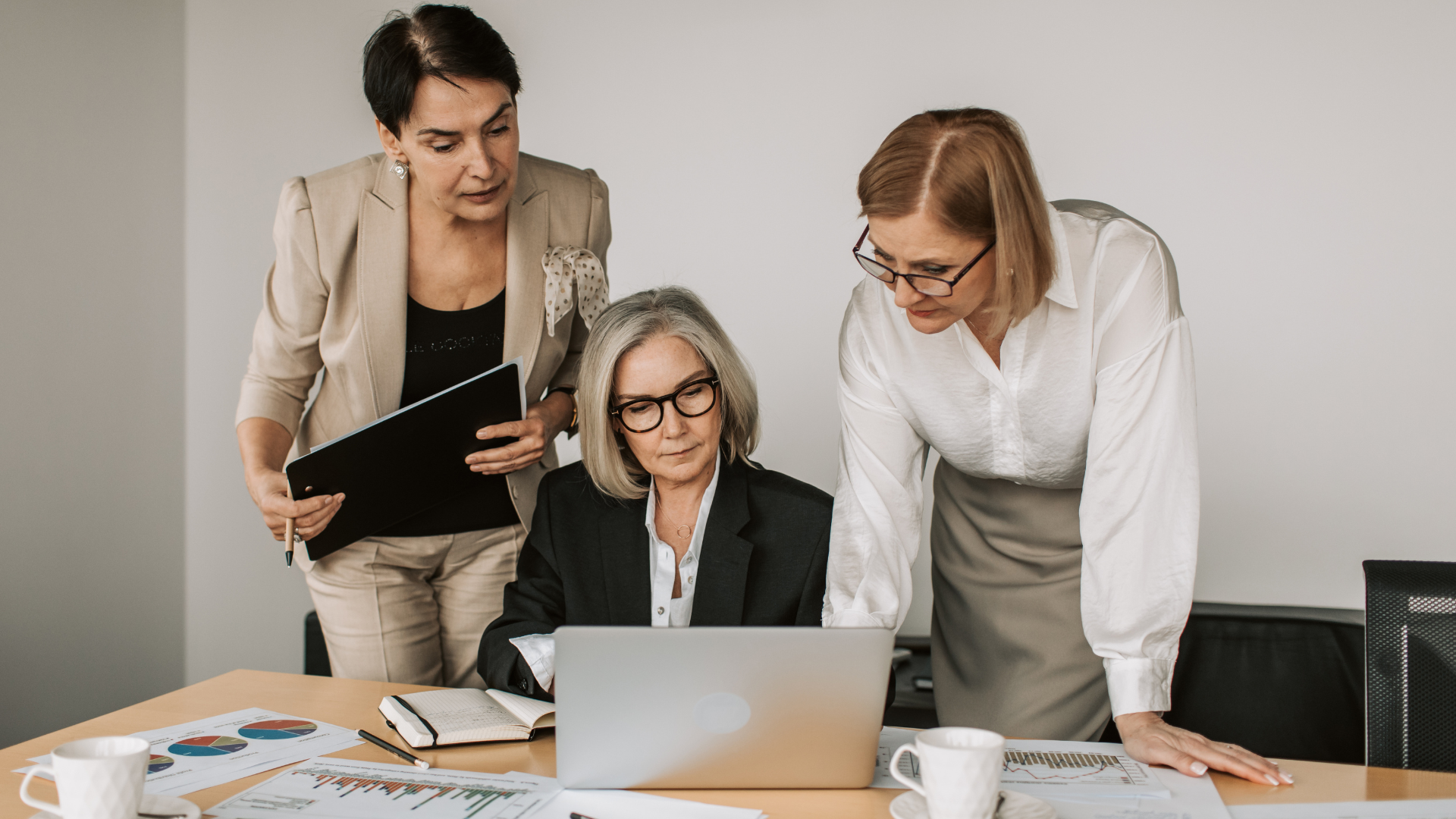 Three women at a table looking at a laptop. Two stand, one leans in; one is seated. Office setting.