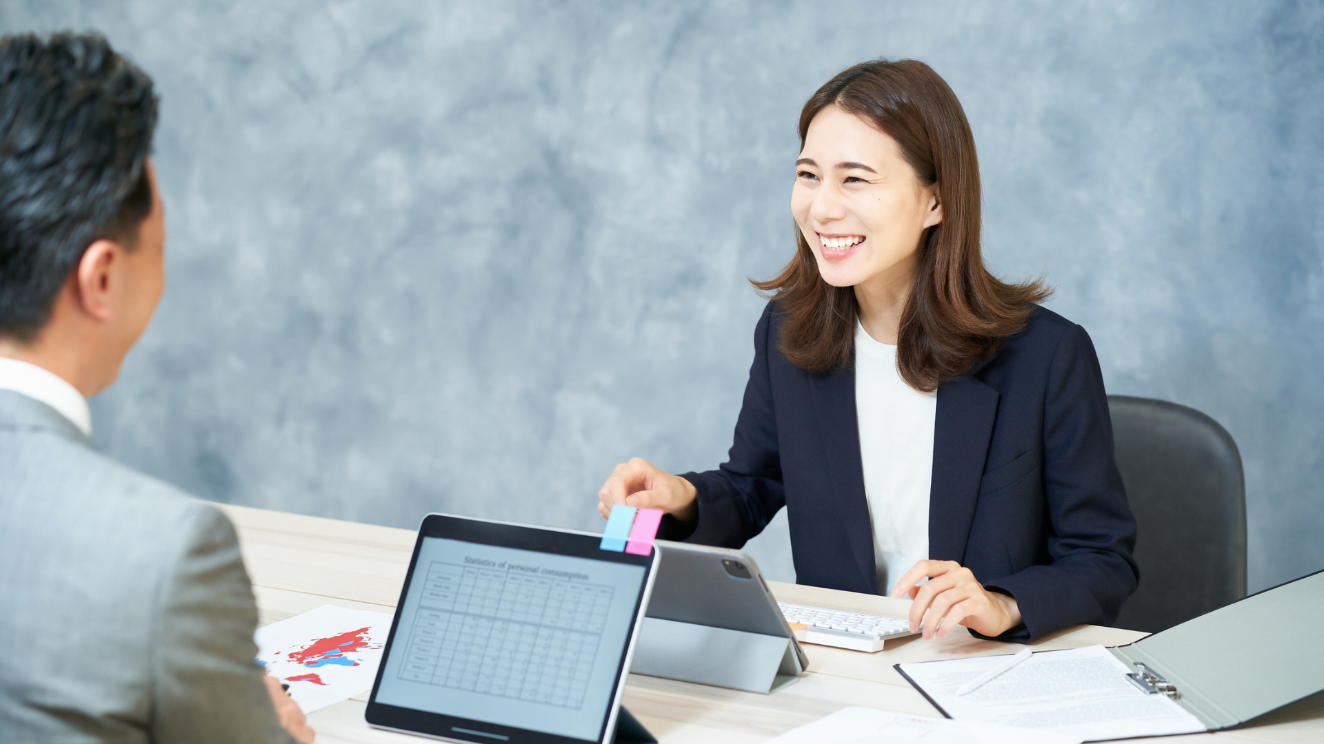 Woman in a navy blazer smiles while speaking with a person at a desk. Tablets and papers are present.