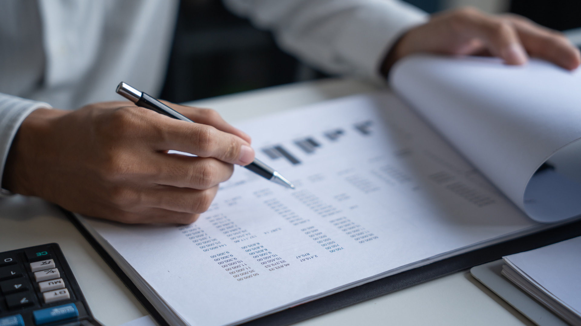 Person reviewing financial documents with a pen; calculator visible.