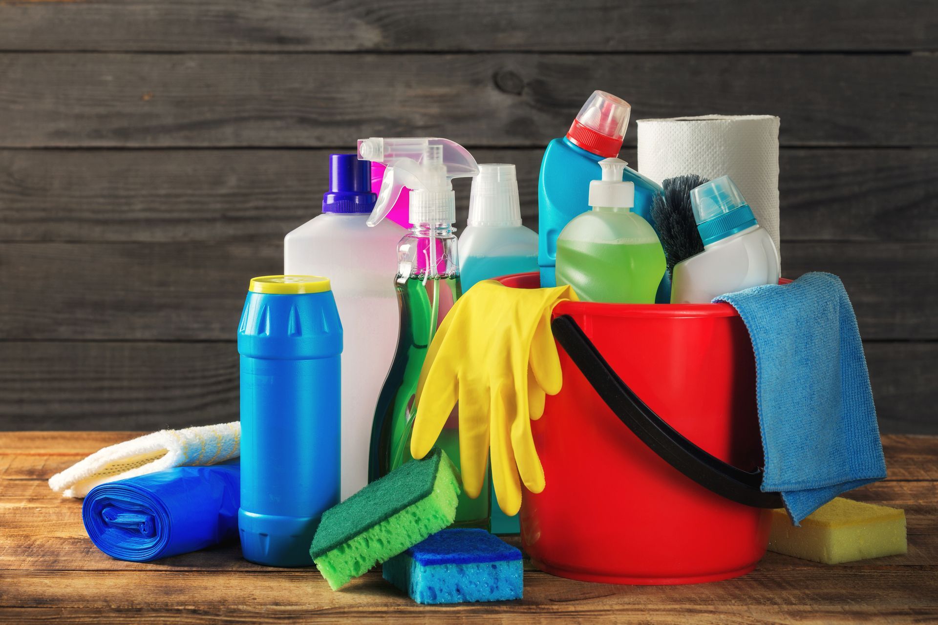 Cleaning supplies on a wooden surface: spray bottles, sponges, gloves, bucket, and paper towels.