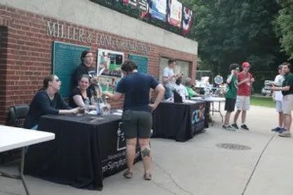 A group of people are standing around tables outside the Big Train baseball field campus.