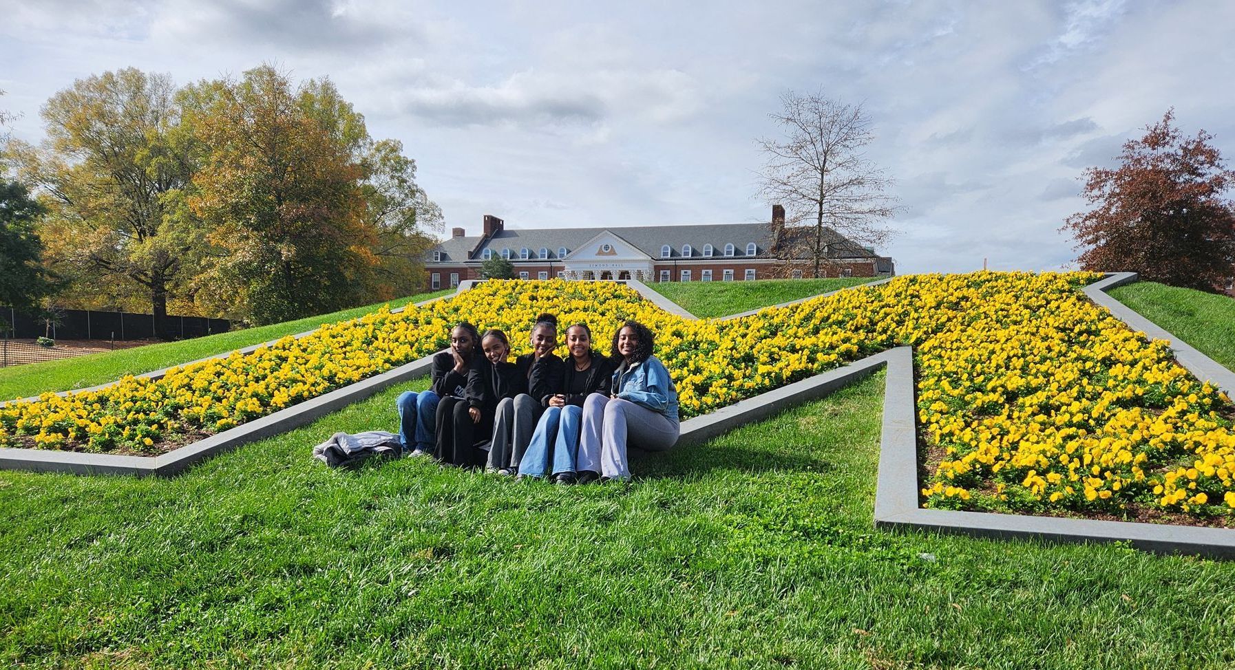 A group of teens are sitting in the grass in front of a yellow flowered University of Maryland M.