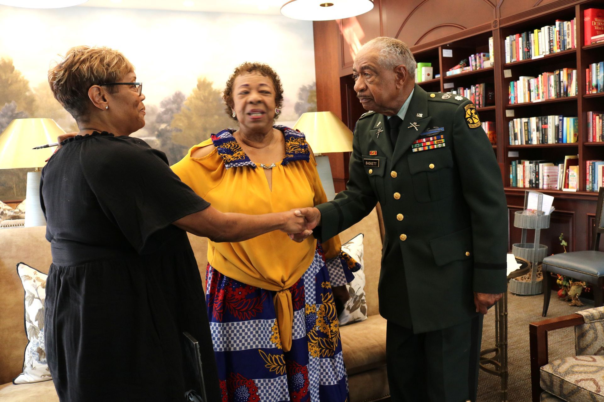 Two women and a man in a military uniform shake hands in a room with a bookshelf and a painting.