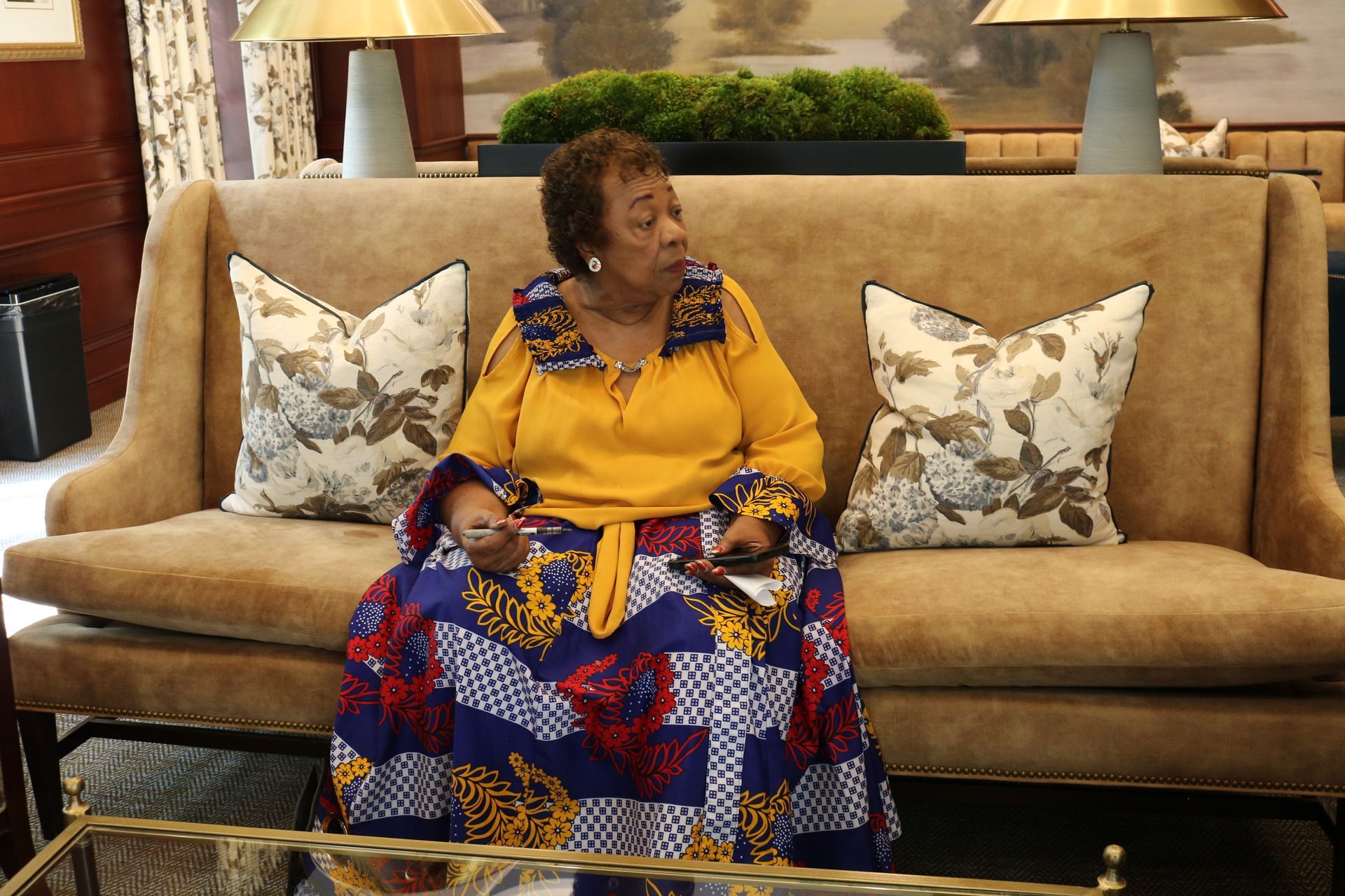 Woman in yellow blouse and patterned skirt sits on a brown sofa in a well-lit room.