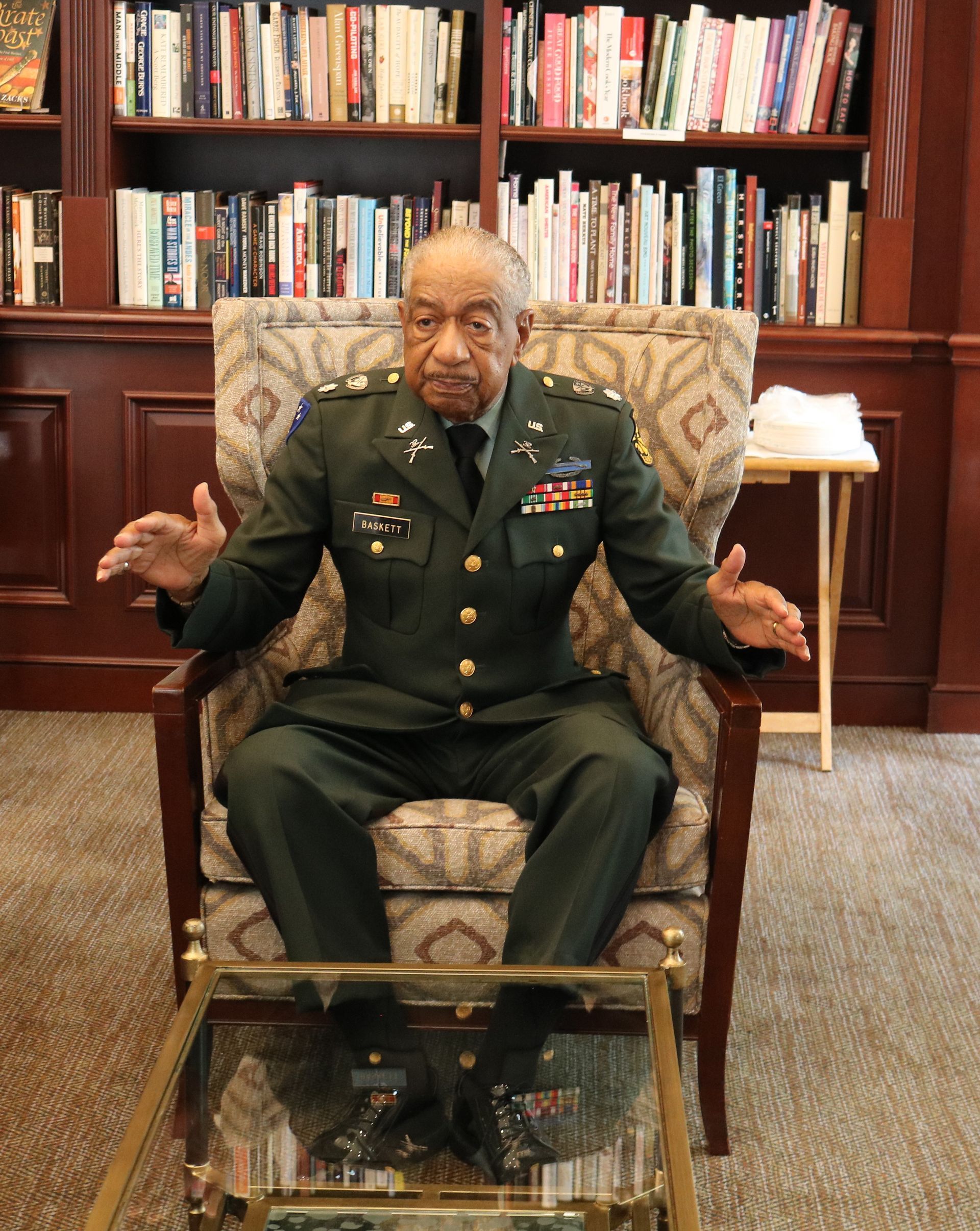 Man in military uniform seated, gesturing with hands in front of a glass table; bookshelves in background.