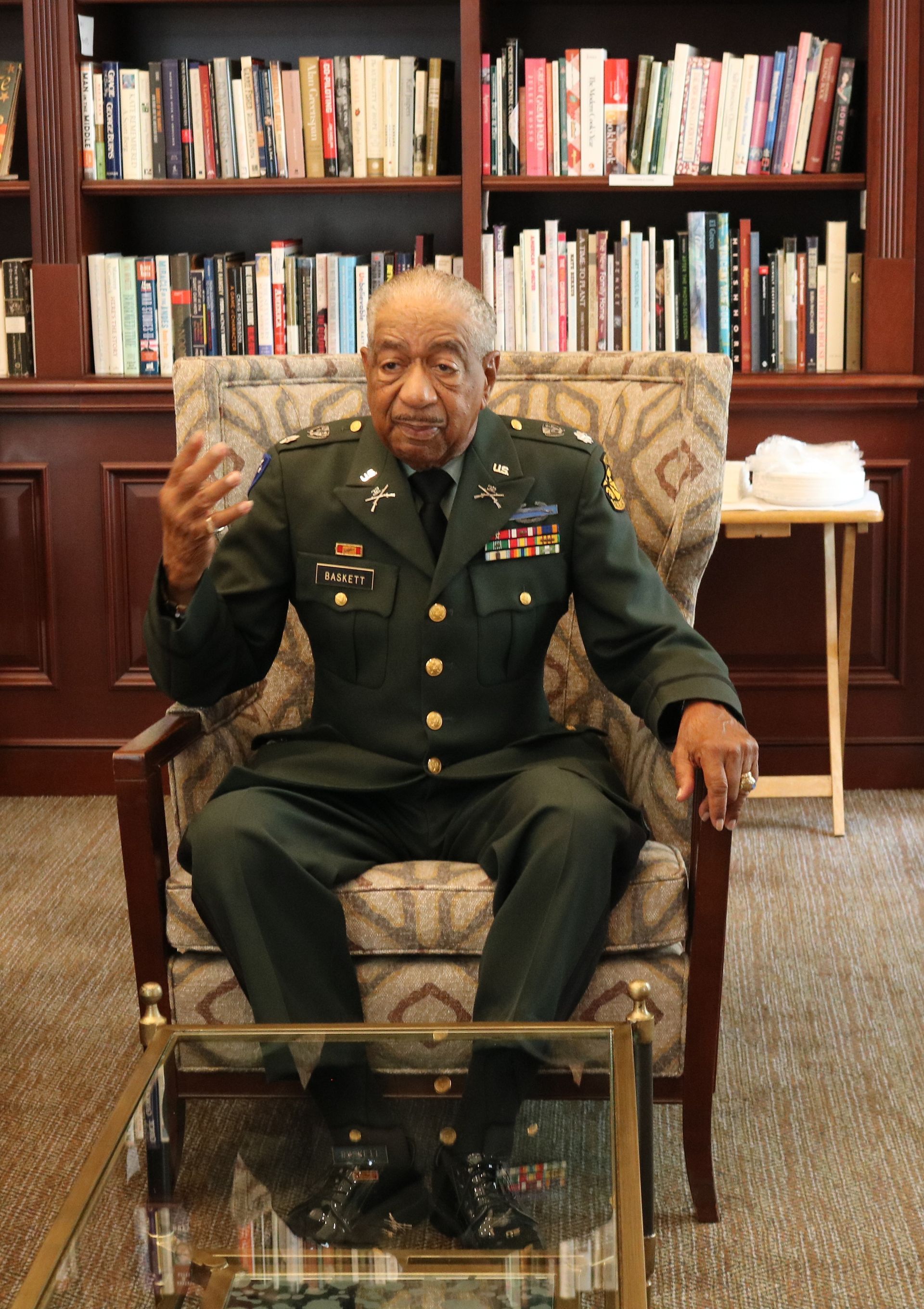 Man in military uniform seated, gesturing. Bookshelves in background.
