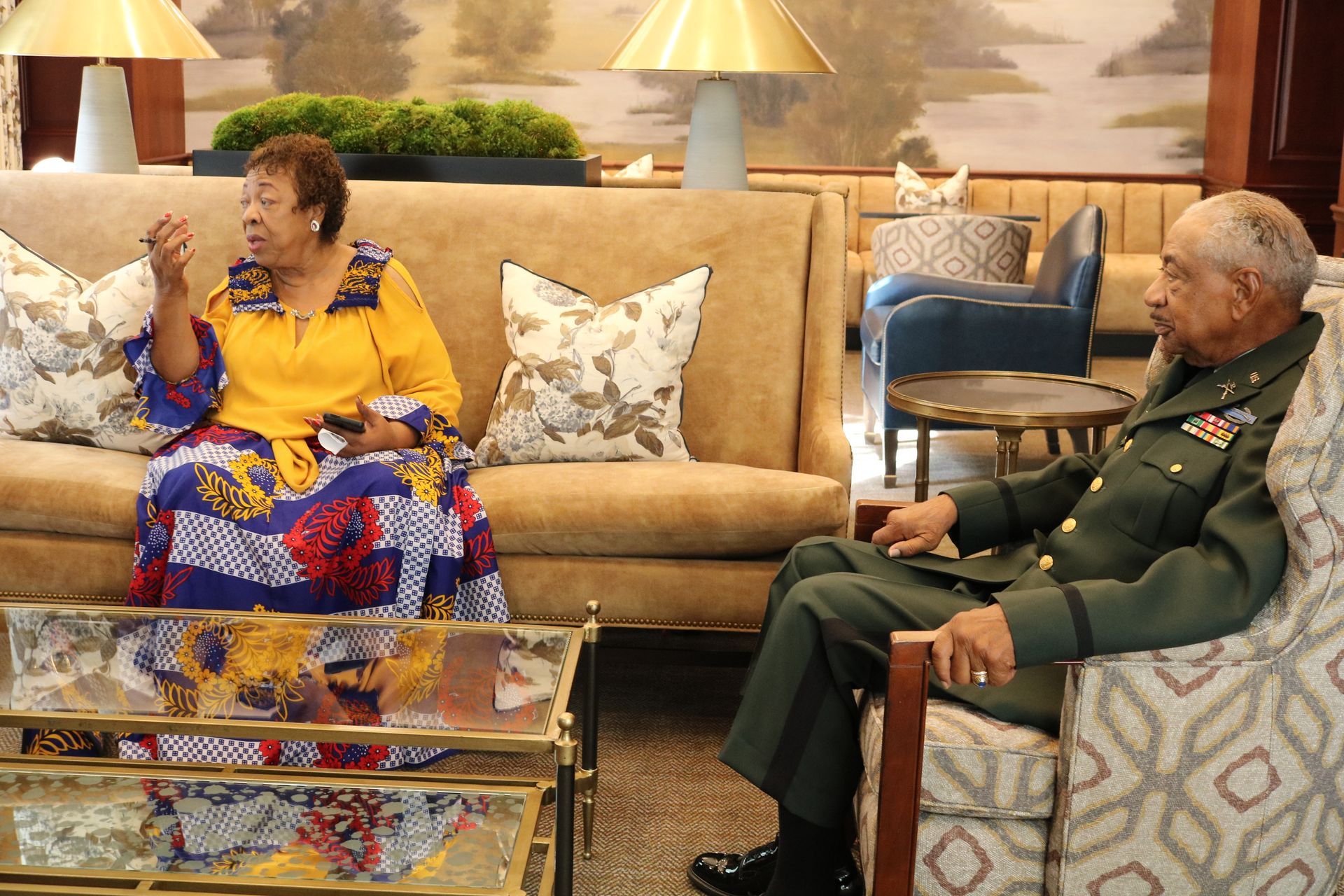 Woman in yellow dress and man in military uniform seated, conversing in a well-lit room with sofa and gold accents.