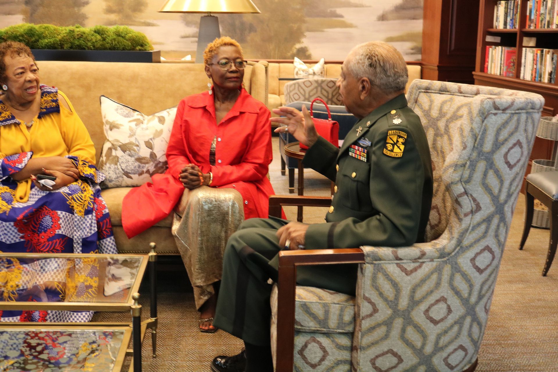 Three people seated, talking. Man in military uniform gesturing, two women listen; indoors.