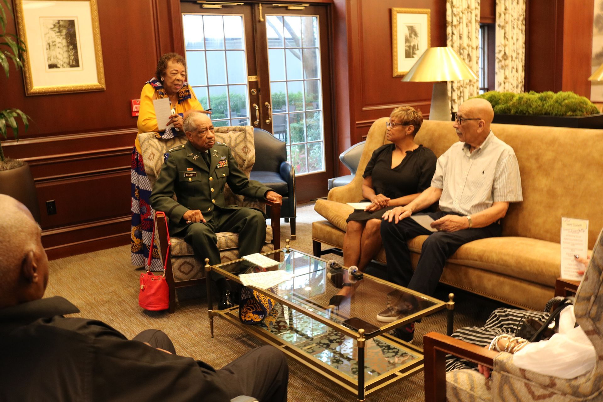 People in a lounge setting, some seated on couches, others standing. One person in a military uniform.
