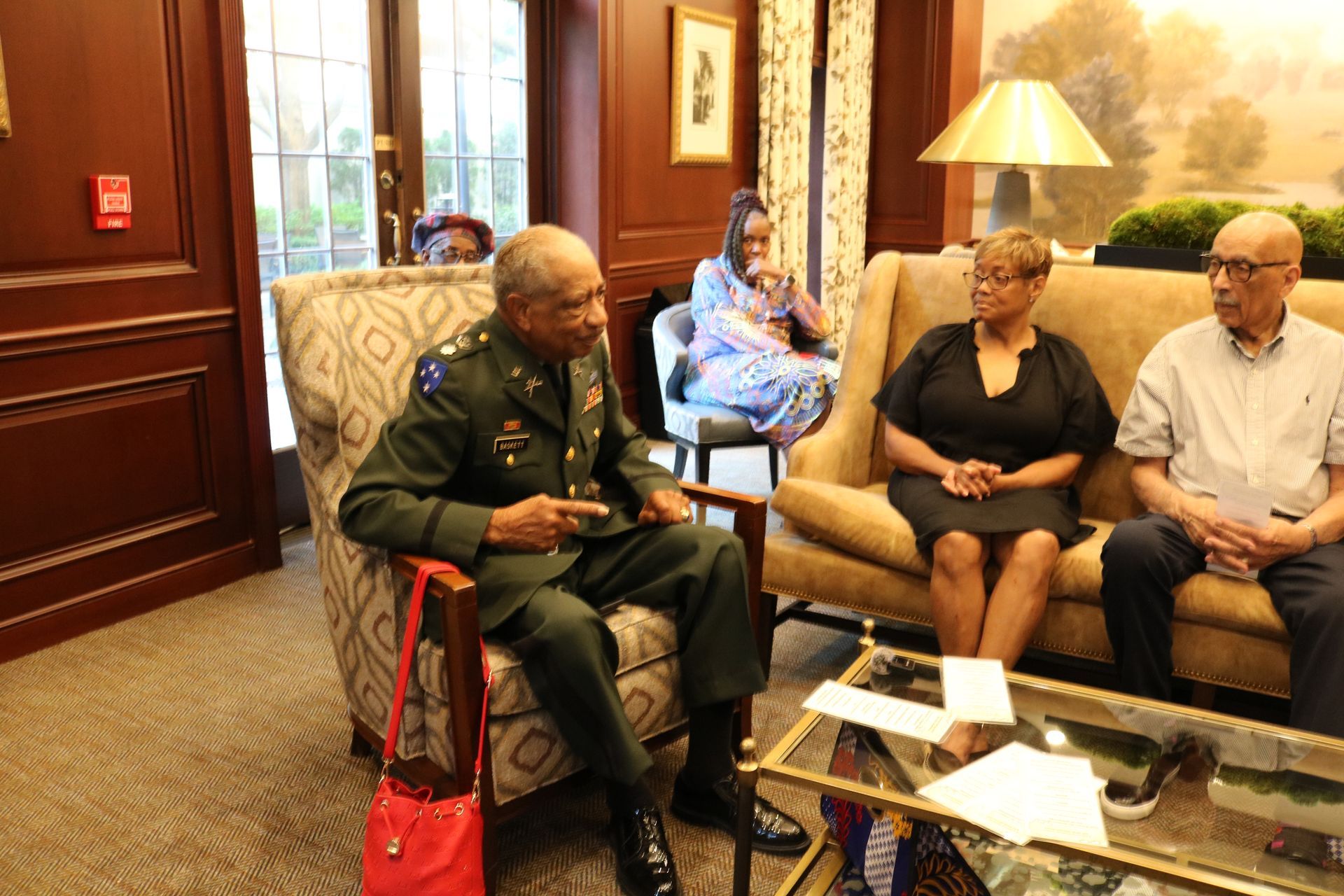 Man in military uniform seated, speaking to two people on a couch. Another person sits nearby. Interior setting.