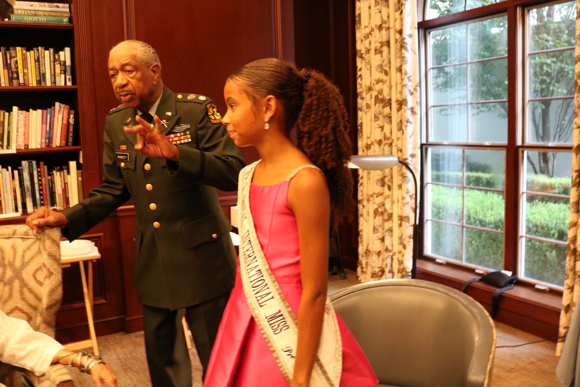 Man in uniform gestures, standing with girl in pink dress and sash indoors near bookshelves and a window.