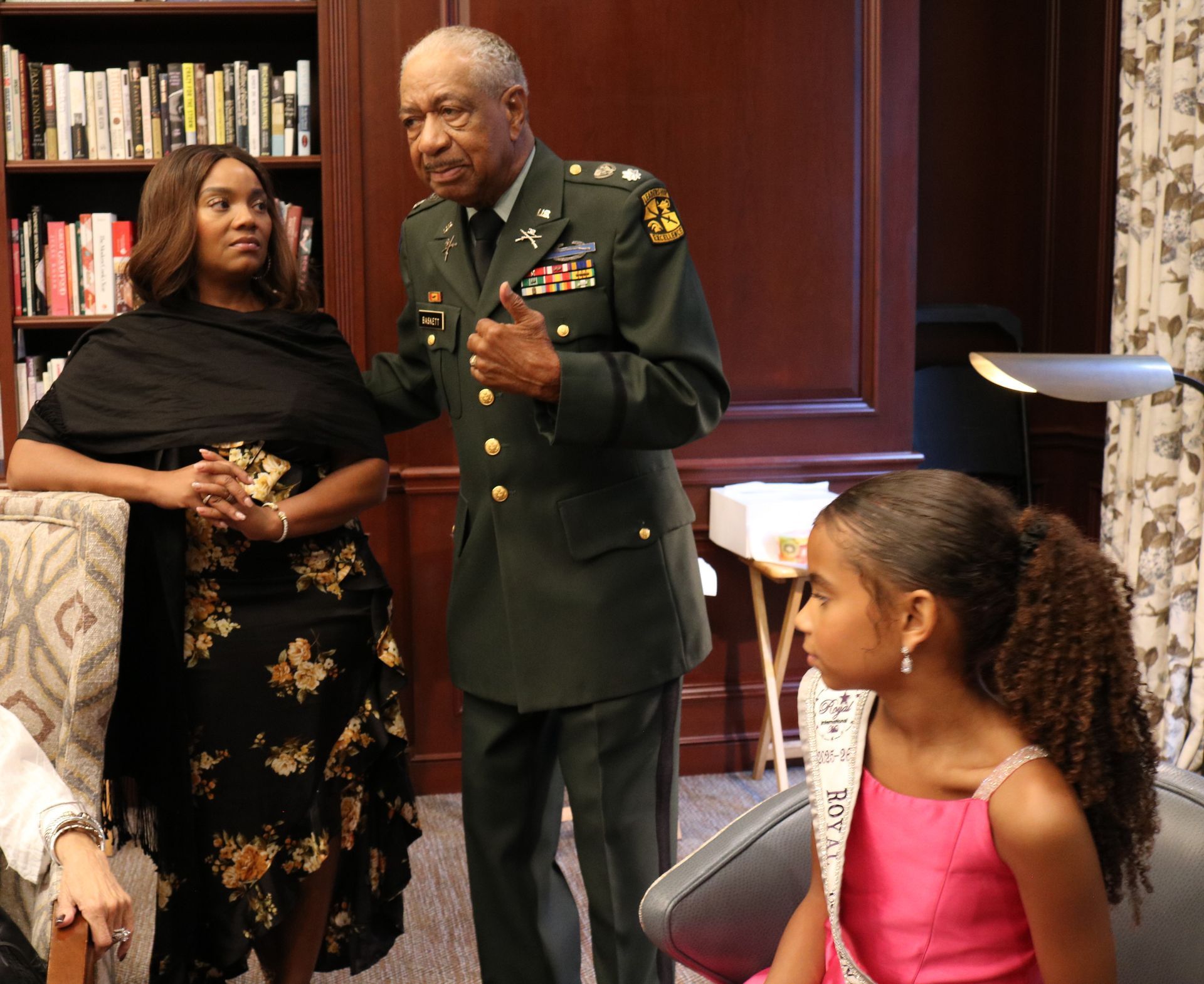 Man in military uniform speaks to a woman and girl, bookcase background.