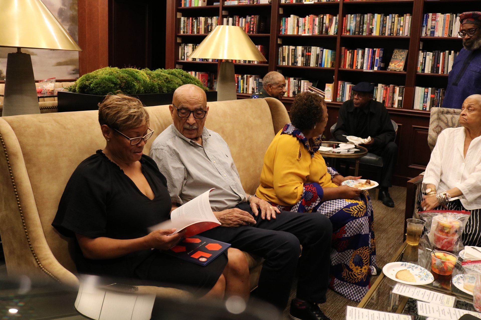 People seated and gathered in a library setting, some reading, others eating. Bookshelves in background.