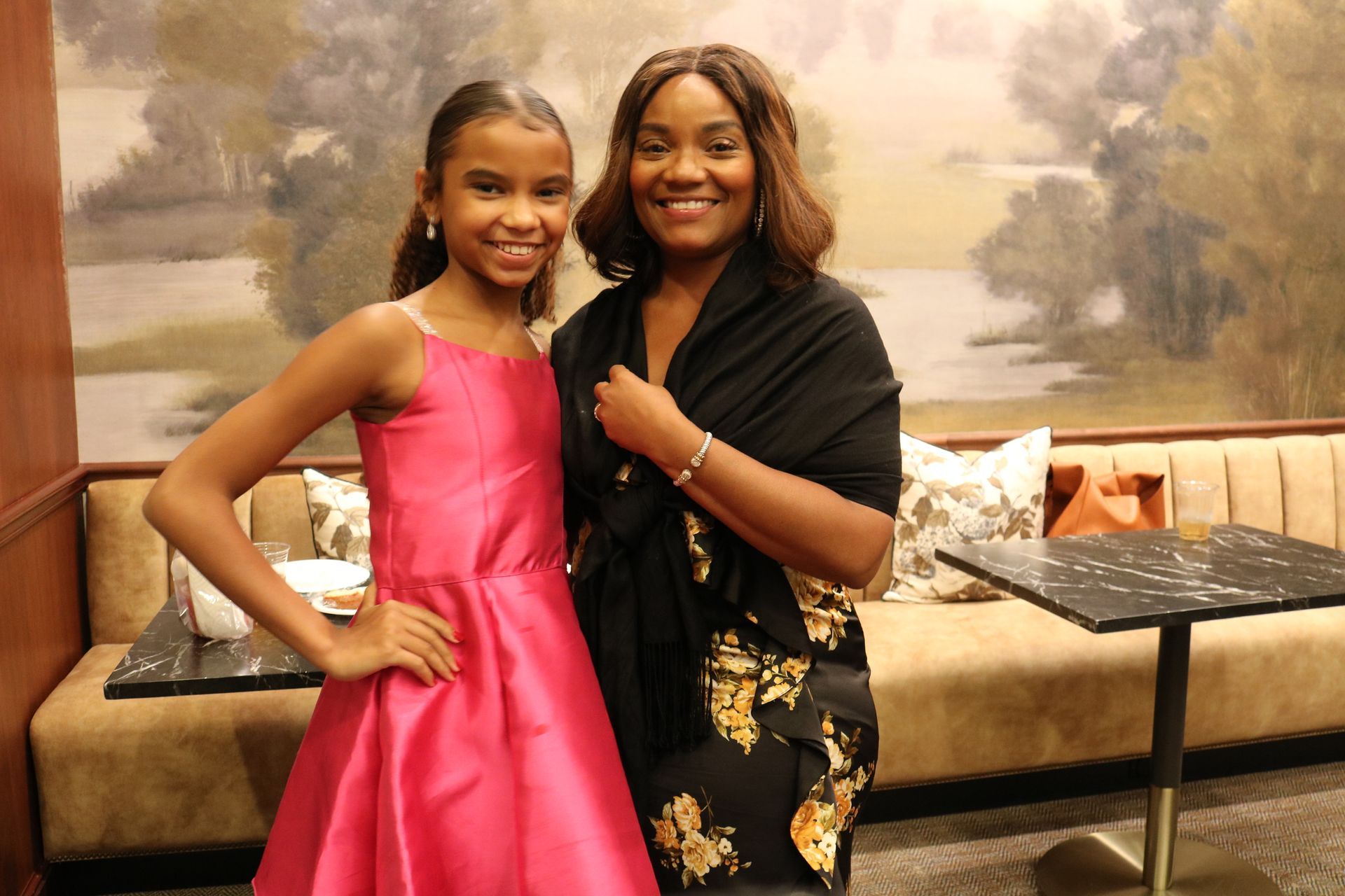 Woman and girl smiling in a room. Girl in pink dress, woman in black shawl and dress. Neutral background.