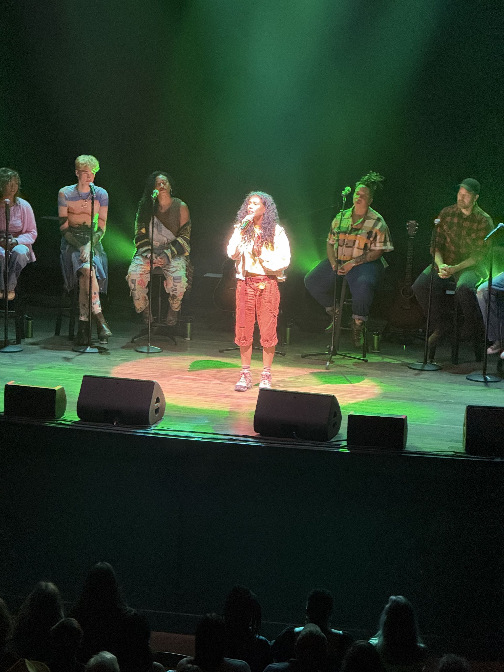 A performer sings on stage, surrounded by seated individuals, under green lighting.