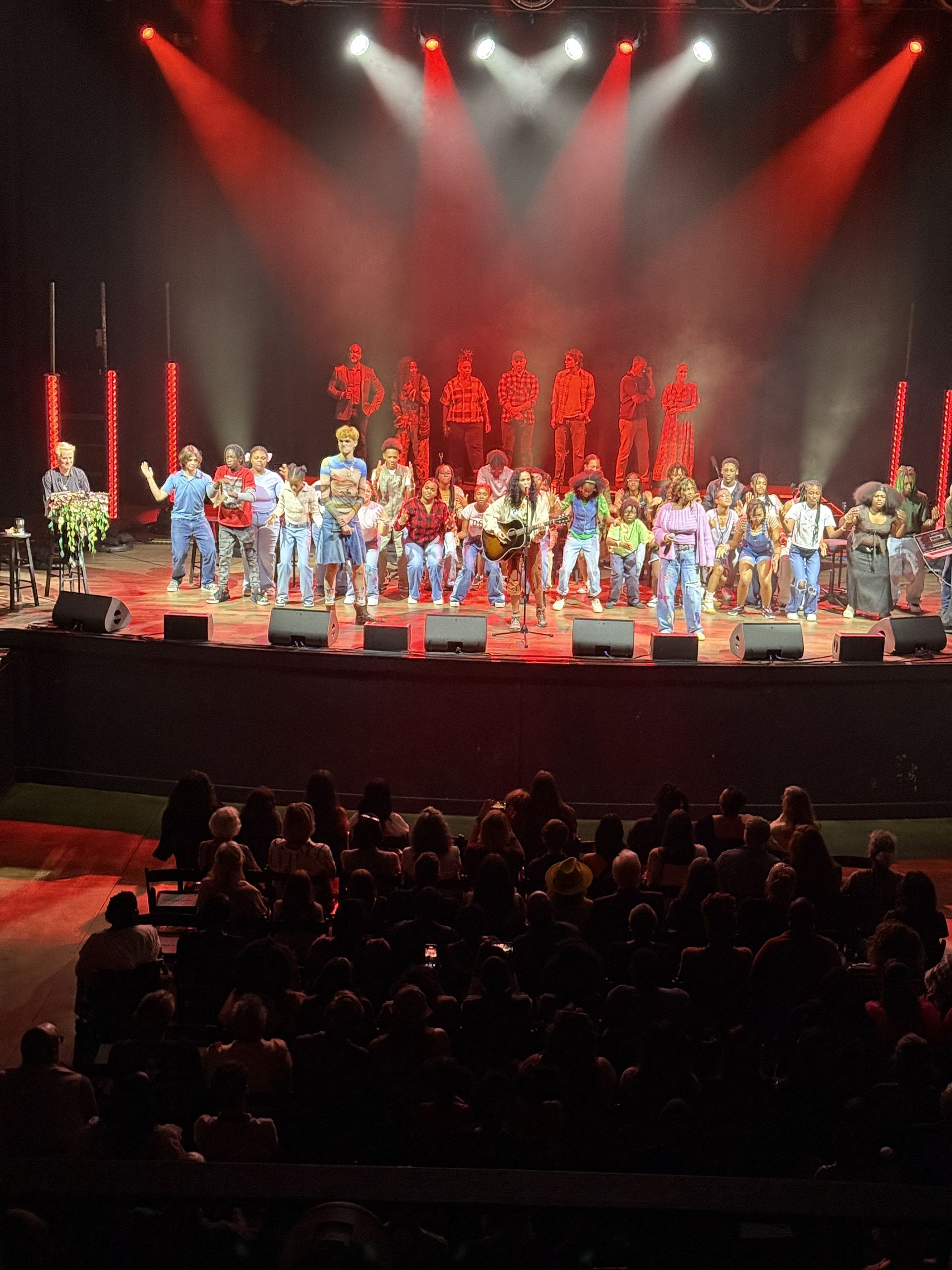 Large group of performers on stage under red lights, facing an audience.