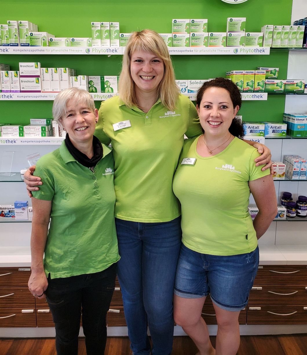 Drei Frauen in grünen Hemden lächeln in einer Apotheke, im Hintergrund sind Regale mit Produkten zu sehen.