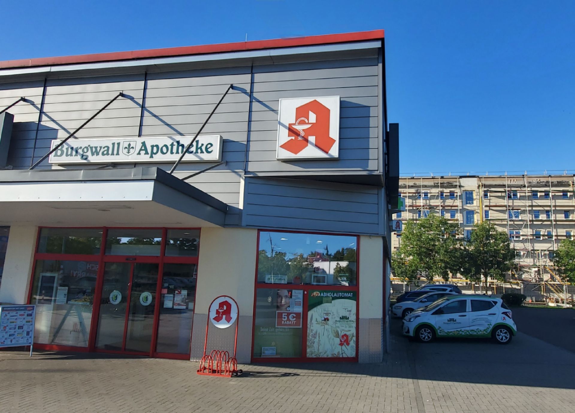 Eine Apotheke mit einem roten „A“-Logo. Im Hintergrund sind Autos und ein großes Wohnhaus zu sehen.